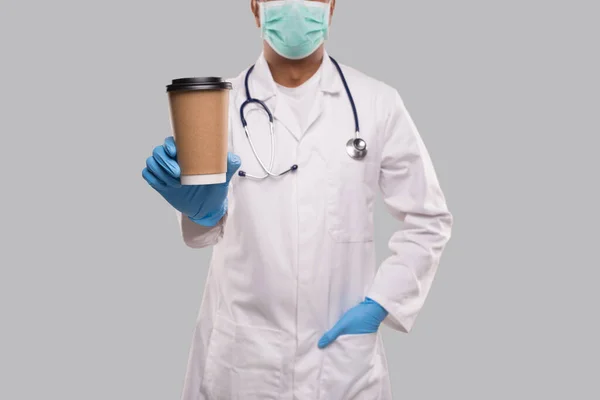Hintli Doktor, tıbbi maske ve eldiven takıp kahve kupası gösteriyor. Hintli doktor kahve tutuyor.. — Stok fotoğraf