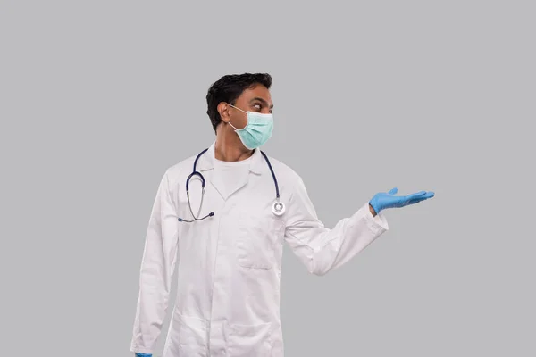 Doctor sosteniendo mano a lado usando máscara médica y guantes mirando a un lado aislado. indio hombre médico signo —  Fotos de Stock