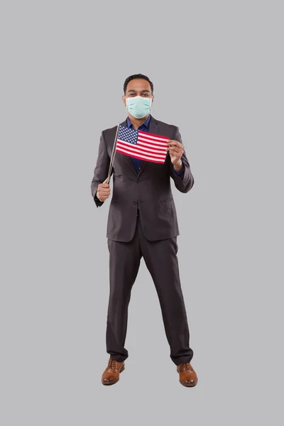 Homem de negócios segurando bandeira americana vestindo máscara médica. Empresário indiano de pé de comprimento total com bandeira dos EUA em mãos — Fotografia de Stock