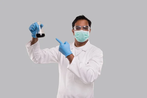 Mannelijke Doctor Pointing op Flask met kleurrijke vloeibare dragen medische masker, handschoenen en glazen geïsoleerd. Wetenschap, Medisch, Virusconcept. Indiase man dokter glimlachen. — Stockfoto