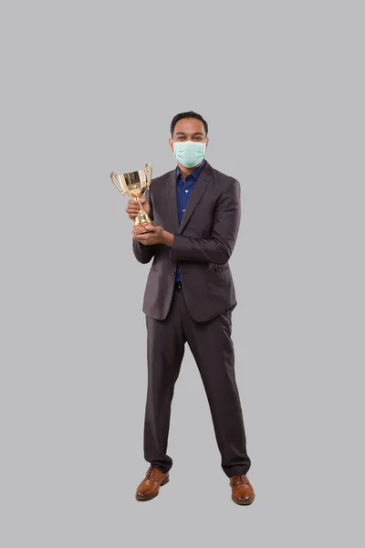 Zakenman met een trofee die een medisch masker draagt. Indiase zakenman staande volle lengte met trofee in handen — Stockfoto