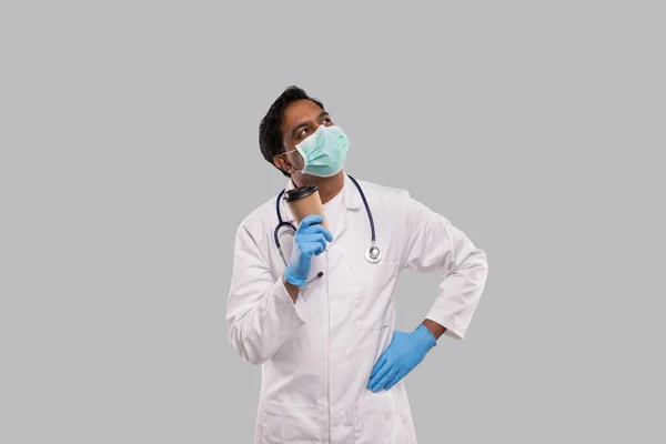 Indian Man Doctor Holding Coffee Take Away Cup dragen van medische maskers en handschoenen denken Kijken omhoog geïsoleerd. Indiase arts houdt koffie te gaan Cup. — Stockfoto