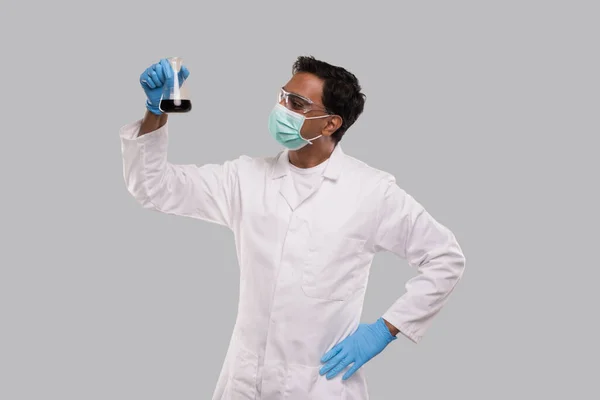 Masculino Doctor Examing Flask con líquido colorido que usa máscara médica, guantes y gafas aislados. Ciencia, Medicina, Concepto Virus. indio hombre médico molienda . — Foto de Stock