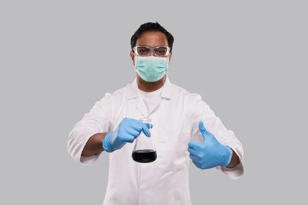 Man Doctor Holding Flask met kleurrijke vloeibare weergegeven duim omhoog dragen van medische masker, handschoenen en glazen geïsoleerd. Wetenschap, Medisch, Virusconcept. Indiase man dokter glimlachen. — Stockfoto