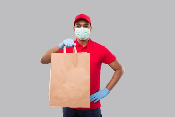 Delivery Man With Paper Bag in Hands WEaring Medical Mask and Gloves Isolated. Red Uniform Indian Delivery Boy. Home Food Delivery. Paper Bag. Man