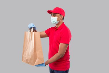 Ellerinde kağıt torba olan teslimatçı tıbbi maske ve eldivenleri tek başına izliyor. Kırmızı üniformalı Kızılderili Teslimatçı Çocuk. Ev yemeği teslimatı. Kağıt Torba Teslimatı