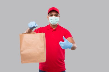 Ellerinde kağıt torba olan teslimatçı, tıbbi maske takıyor ve başparmağını kaldırıyor. Kırmızı üniformalı Kızılderili Teslimatçı Çocuk. Ev yemeği teslimatı. Kağıt Torba Teslimatı