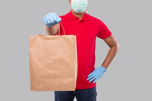 Delivery Man With Paper Bag in Hands Wearing Medical Mask and Gloves Isolated Close Up. Red Uniform Indian Delivery Boy. Home Food Delivery. Paper Bag Delivery