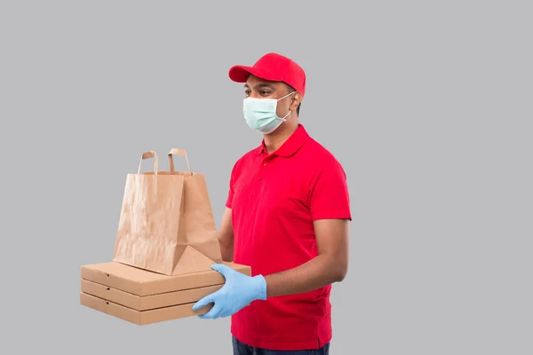 Delivery Man With Paper Bag and Three Pizza Box in Hands Wearing Medical Mask and Gloves Watching Side Isolated. Red Uniform Indian Delivery Boy. Home Food Delivery. Paper Bag Delivery