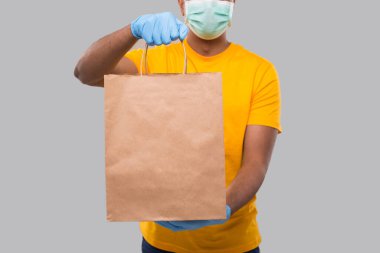 Ellerinde kağıt torba olan teslimatçı tıbbi maske ve eldiven takıyor. Kapalı. Sarı üniformalı Kızılderili Teslimatçı Çocuk. Ev yemeği teslimatı. Kağıt Torba