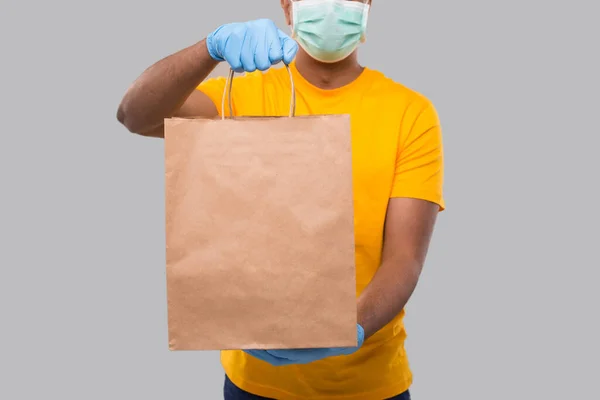 Ellerinde kağıt torba olan teslimatçı tıbbi maske ve eldiven takıyor. Kapalı. Sarı üniformalı Kızılderili Teslimatçı Çocuk. Ev yemeği teslimatı. Kağıt Torba — Stok fotoğraf
