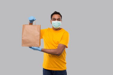 Ellerinde kağıt torba olan teslimatçı tıbbi maske ve eldivenler takıyor. Sarı üniformalı Kızılderili Teslimatçı Çocuk. Ev yemeği teslimatı. Kağıt Torba