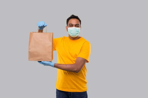 Ellerinde kağıt torba olan teslimatçı tıbbi maske ve eldivenler takıyor. Sarı üniformalı Kızılderili Teslimatçı Çocuk. Ev yemeği teslimatı. Kağıt Torba — Stok fotoğraf