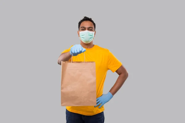 Szállítási ember papírzacskóval a kezében visel orvosi maszk és kesztyű elszigetelt. Sárga egyenruhás indiai kifutófiú. Házhozszállítás. Papírzacskó — Stock Fotó
