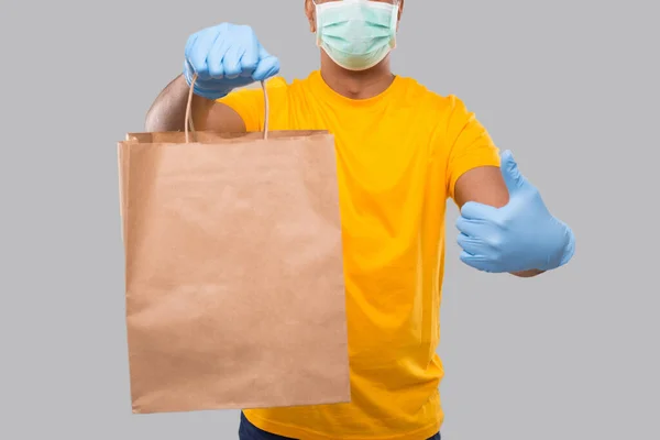 Delivery Man With Paper Bag in Hands Wearing Medical Mask and Gloves Visar tummen upp Isolerad närbild. Gul enhetlig indisk springpojke. Hem Matleveranser. Papperspåse — Stockfoto