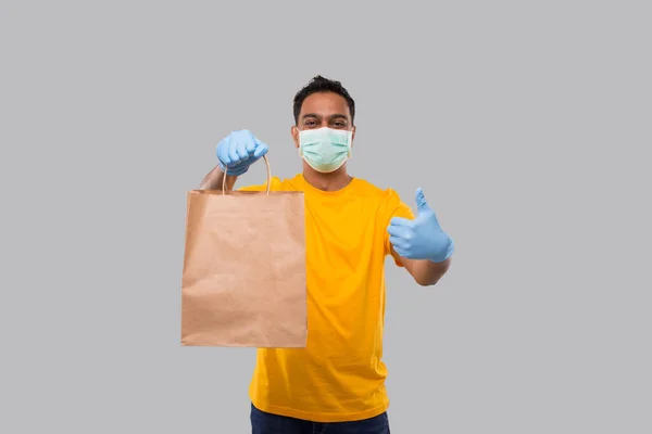 Liefermann mit Papiertüte in der Hand, mit medizinischer Maske und Handschuhen, die isoliert den Daumen nach oben zeigen. Gelbe Uniform indischer Lieferjunge. Essenslieferung nach Hause. Papiertüte — Stockfoto