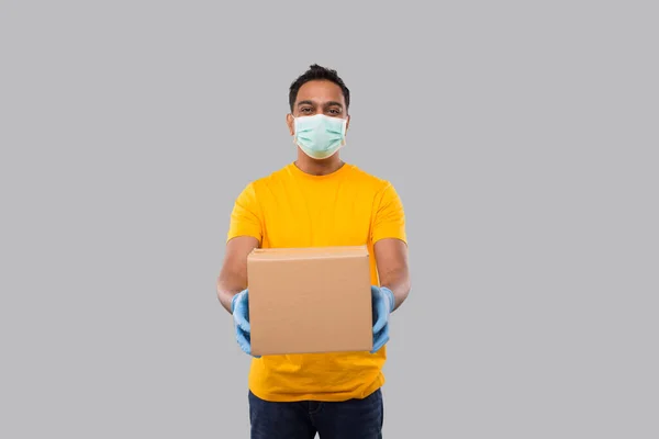 Hombre indio con caja en las manos con máscara médica y guantes aislados. Camiseta amarilla Delivery Boy. Entrega a domicilio. Héroe de cuarentena. Hombre sonriendo —  Fotos de Stock