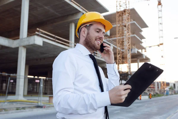Construtor de tomada no telefone assistindo Clipboard. Empresário vestindo Hardhat perto de Obras de Construção. Edifício — Fotografia de Stock