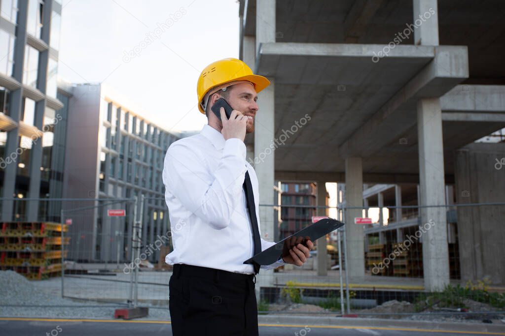 Builder Taking on Phone watching Clipboard. Businessman wearing Hardhat near Construction Works. House Building