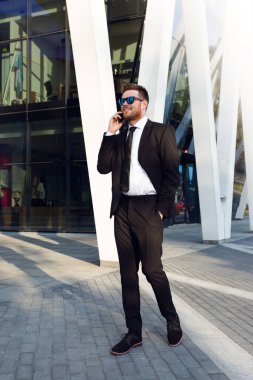 İş adamı telefonda konuşurken izole edildi. İş adamı elinde telefonla tam yol ayakta duruyor. Güneşli bir günde iş adamı. Güneş gözlüğü takıp telefonda konuşuyor.