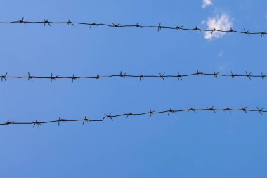 Arkadaki mavi gökyüzü ve beyaz bulutların dikenli telleri. Sınır, hapishane ya da göç kavramı. Üç dikenli tel. Çitli bölgenin dikenli telleri. Gizli nesne sınırı, kapalı engel.