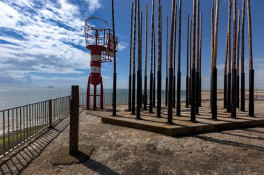 Vlissingen - Badstrand sahilindeki rüzgar orgu yanında beyaz-kırmızı deniz feneri. Arka planda mavi deniz ve gemi..