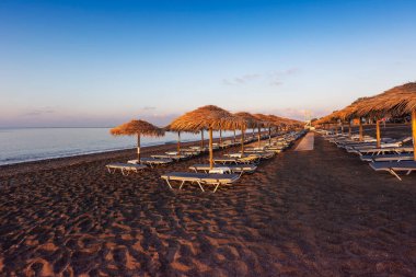 Perissa, Perivolos, Santorini siyah plajı. Güneşin sabah ışıklarıyla aydınlatılıyor. Arka arkaya dizilmiş şemsiyeli plaj sandalyeleri. Güzel sakin deniz.
