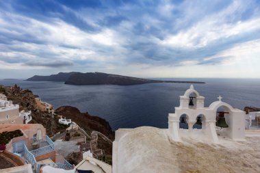 Santorini, Oia, uçurumdan kalderaya bak. Sağda beyaz çan kulesi, solda beyaz çatı Yunan beyaz evlerinin uçurumunda. Arka planda bir ada ve dramatik bulutlar.