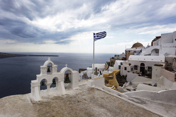 Santorini - Oia, view from the cliff to the caldera. White bell tower on the right and a white roof on the left on the cliff of Greek white houses. In the background an island and dramatic clouds.