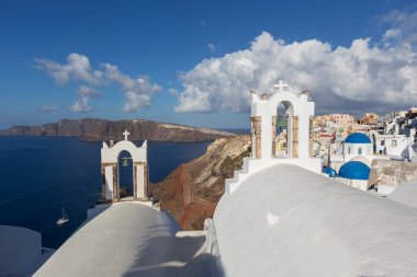 Santorini, Oia - beyaz iki çan kulesinin manzarası, arka planda mavi kubbeli bir kilise ve denizin üstünde beyaz bulutlu güzel mavi gökyüzü.