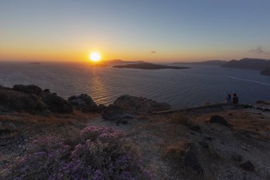 Santorini Caldera 'nın üzerinde güzel bir günbatımı, kayalıklarda kayalar ve arka planda batan güneşle birlikte tüm sahili aydınlatan çiçekli bir fundalığın önünde..