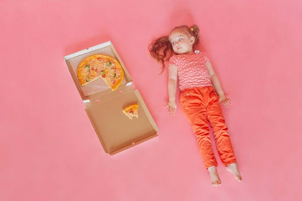 Küçük tatlı mutlu kız pembe bir arka planda pizzanın yanında bir kutuda yatıyor ve ona bakıyor. Boşluğu kopyala