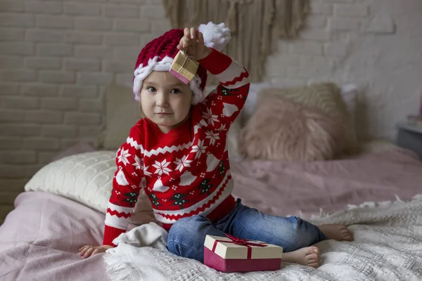 Kırmızı Noel kazaklı küçük kız ve Noel Baba şapkası hediyelerle yatakta oynuyor.