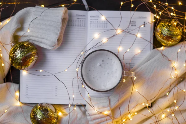 Yeni yıl ya da Noel kış kompozisyonu. Sıcak bir süveter, Noel ışıkları, not defteri ve kahveyle mevsimlik Noel havası. Kopyala lütfen. Seçici odak