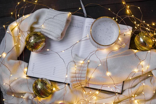 Yeni yıl ya da Noel kış kompozisyonu. Sıcak bir süveter, Noel ışıkları, not defteri ve kahveyle mevsimlik Noel havası. Kopyala lütfen. Seçici odak