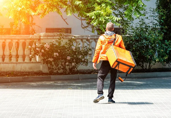 Delivery man with backpack. caucasian deliveryman isolated in orange uniform on park background. Contacless delivery service hurrying up during quarantine. Man delivers food during isolation. Safety.