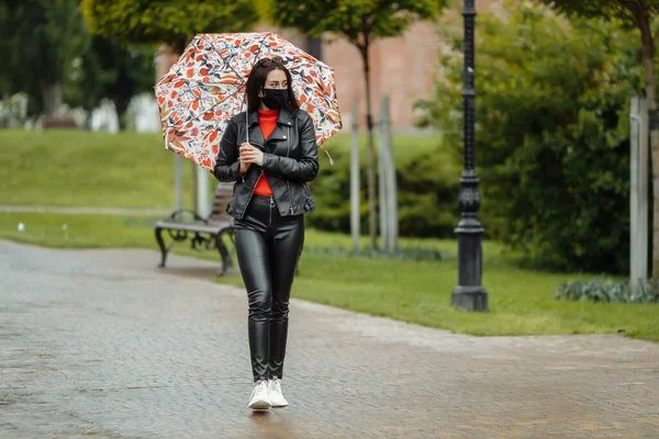 Ein maskiertes Mädchen läuft die Straße entlang. Ein Mädchen mit Schutzmaske geht mit einem Regenschirm im Regen durch den Park. Coronavirus-Infektion COVID-19 — Stockfoto