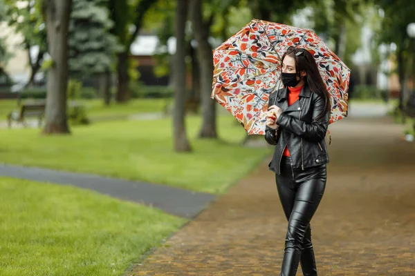 Uma menina mascarada está andando pela rua. Uma menina com uma máscara protetora caminha no parque com um guarda-chuva na chuva. Infecção pelo vírus da coronária COVID-19 — Fotografia de Stock