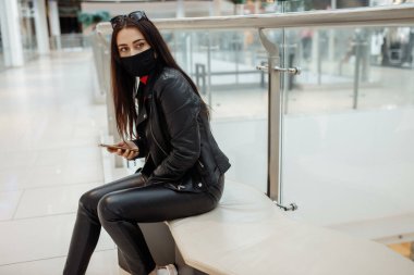 Alışveriş merkezinde siyah maskeli ve cep telefonu olan bir kız. Koronavirüs salgını. Maskeli bir kadın alışveriş merkezinde duruyor. Koruyucu maskeli bir kız alışveriş merkezinde alışveriş yapıyor.