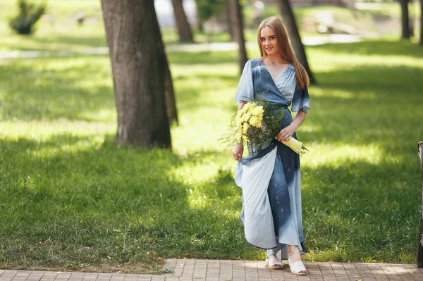 Stylowa dziewczyna pozowanie w sukience w słonecznym parku wiosennym. Spokojny portret pięknej dziewczyny stojącej z bukietem wiosną — Zdjęcie stockowe