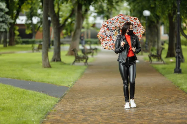 Uma menina mascarada está andando pela rua. Uma menina com uma máscara protetora caminha no parque com um guarda-chuva na chuva. Infecção pelo vírus da coronária COVID-19 — Fotografia de Stock