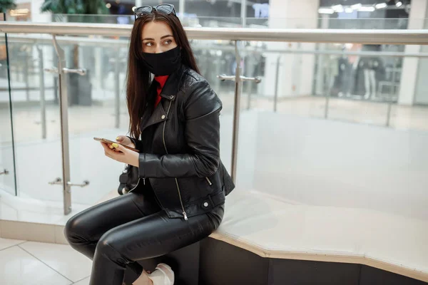 Menina com máscara preta médica e telefone celular em um centro comercial. pandemia de coronavírus. Uma mulher com uma máscara está de pé em um centro comercial. Uma garota com uma máscara protetora está comprando no shopping — Fotografia de Stock