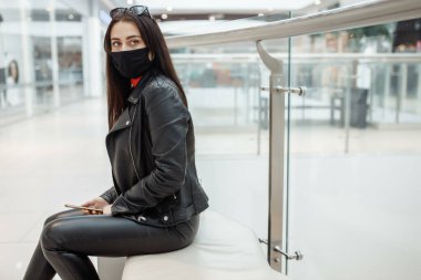 Alışveriş merkezinde siyah maskeli ve cep telefonu olan bir kız. Koronavirüs salgını. Maskeli bir kadın alışveriş merkezinde duruyor. Koruyucu maskeli bir kız alışveriş merkezinde alışveriş yapıyor.