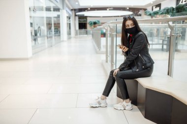 Alışveriş merkezinde siyah maskeli ve cep telefonu olan bir kız. Koronavirüs salgını. Maskeli bir kadın alışveriş merkezinde duruyor. Koruyucu maskeli bir kız alışveriş merkezinde alışveriş yapıyor.