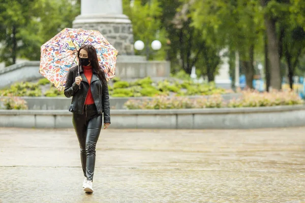 Uma menina mascarada está andando pela rua. Uma menina com uma máscara protetora caminha no parque com um guarda-chuva na chuva. Infecção pelo vírus da coronária COVID-19 — Fotografia de Stock