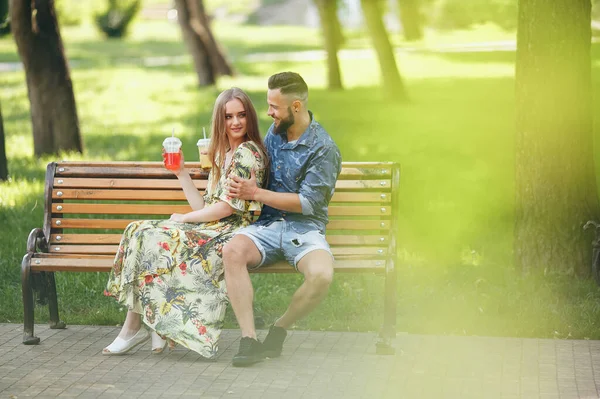 Fashion young teenage couple resting in a city park with drinks sitting on a bench on a sunny summer day — Stock Photo, Image