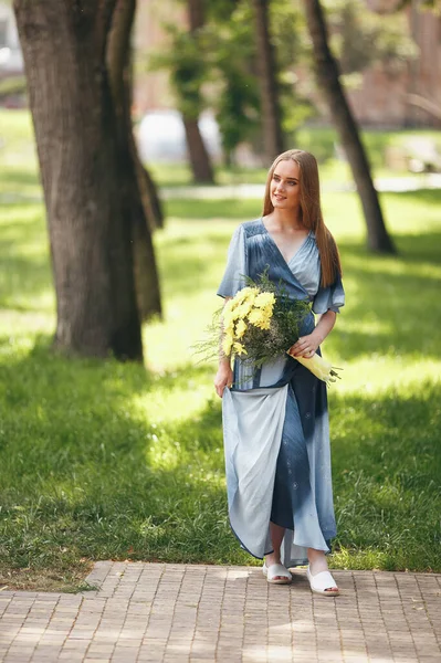 Güneşli bir bahar parkında elbise giymiş şık bir kız. Güzel bir kızın sakin portresi. Baharda elinde bir buketle duruyor. — Stok fotoğraf