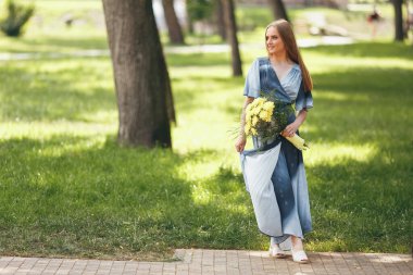 Güneşli bir bahar parkında elbise giymiş şık bir kız. Güzel bir kızın sakin portresi. Baharda elinde bir buketle duruyor.