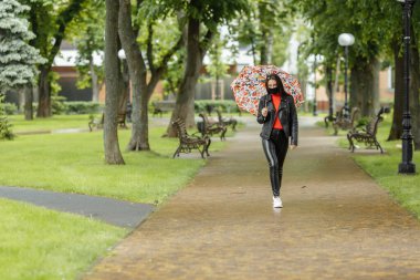 Maskeli bir kız cadde boyunca yürüyor. Koruyucu maskeli bir kız yağmurda şemsiyeyle parkta yürür. Coronavirus enfeksiyonu COVID-19