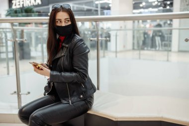 Alışveriş merkezinde siyah maskeli ve cep telefonu olan bir kız. Koronavirüs salgını. Maskeli bir kadın alışveriş merkezinde duruyor. Koruyucu maskeli bir kız alışveriş merkezinde alışveriş yapıyor.